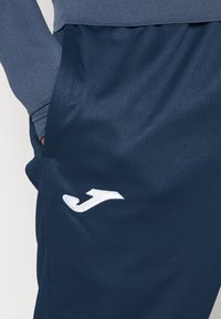 Pantalons de sport bleus marine en tissu lisse, dotés d'une poche latérale et d'un logo brodé blanc sur la cuisse gauche.