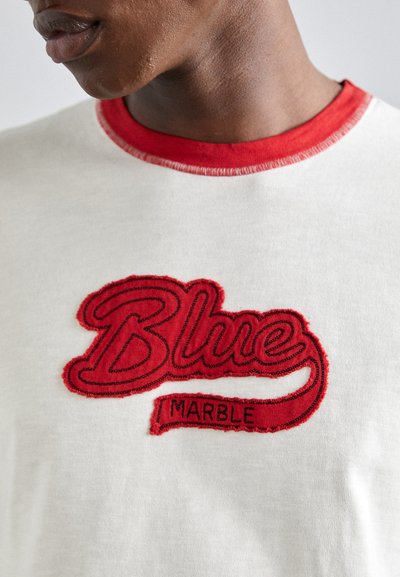 Valge T-särk punase soonikkoha ja silmapaistva punase tikandiga "Blue Marble" logo, millel on tekstuurilt julge fond.