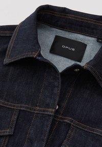 Veste en denim bleu foncé avec des coutures orange contrastantes, un col classique et une étiquette à l'intérieur affichant "OPUS."