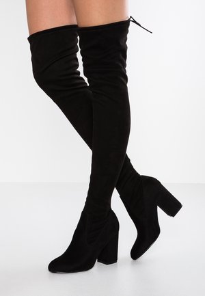 Über dem Knie schwarze Wildlederstiefel mit Blockabsätzen, getragen an übereinandergeschlagenen Beinen vor weißem Hintergrund.