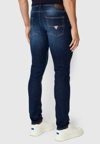 Tummansiniset denim-farkut, joissa on slim fit, taka-taskut ja kontrastivärinen ommel. Yhdistetty valkoisiin tennareihin, mikä korostaa modernia muotoilua.