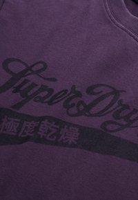 Fioletowa bluza z ciemnym nadrukiem, w tym napisem "SuperDry", wykonana z miękkiej bawełny z klasycznym dekoltem typu crew neck.