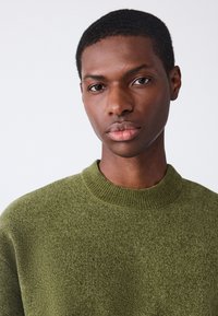 Olivzaļš džemperis ar teksturētu adījumu, ar ribotu apkakli. Dizains ir vienkāršs, bez redzamām rakstu vai akcentu elementiem.