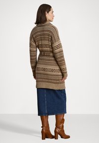 Cardigã de malha em tons de castanho com design estampado, apresentando uma cintura com cinto, punhos canelados e uma silhueta longa e aberta. Combinado com uma saia de ganga e botas castanhas.