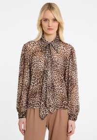 Woman wearing a long-sleeve sheer leopard print blouse with a large pointed collar and matching neck tie, paired with brown pants.