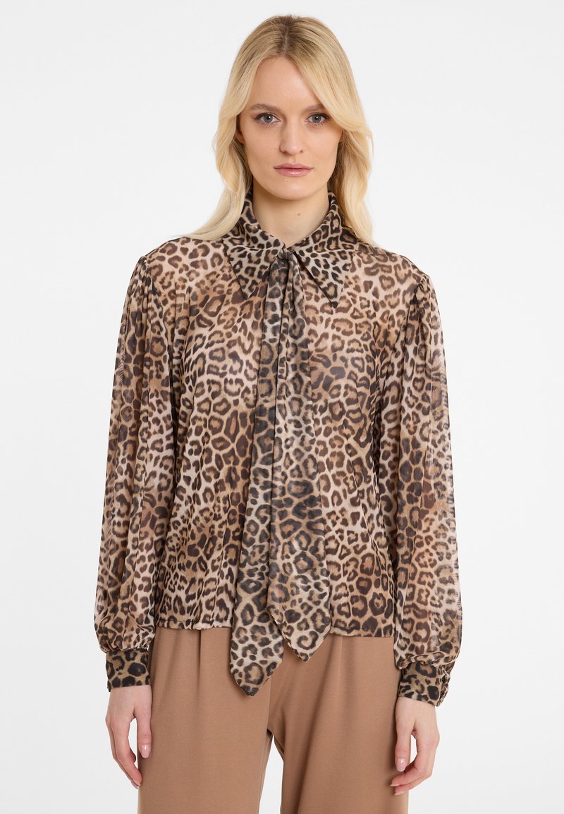 Woman wearing a long-sleeve sheer leopard print blouse with a large pointed collar and matching neck tie, paired with brown pants.