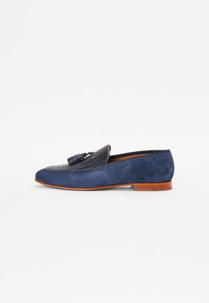 Mocasín azul marino con un panel de cuero trenzado y un adorno de borlas. Cuenta con una parte superior de ante y una suela de cuero con un tacón bajo.