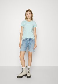 Šviesiai mėlyna trumpomis rankovėmis marškinėliai su balta logotipu, derinami su suveržtais džinsiniais šortais ir béžiniais lace-up batais, turinčiais juodus padus.