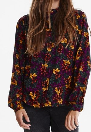 Femme portant un chemisier à manches longues, à motif floral sombre avec des fleurs jaunes, orange et violettes, associé à un pantalon noir.