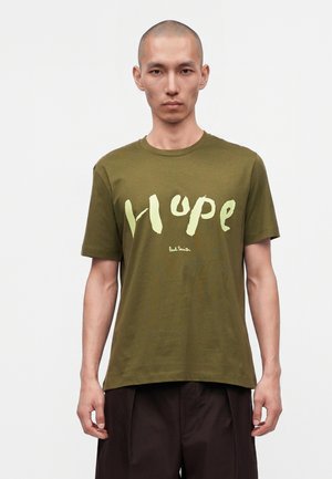 Homme portant un t-shirt vert olive avec de grandes lettres jaune pâle formant le mot « Hope » et un petit texte « Paul Smith » en dessous, debout sur fond blanc.