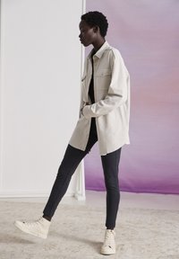 Gai bēšs pārāk liels krekla žakets ar divām krūšu kabatām, apvienots ar melniem pieguļošiem biksēm un krēmkrāsas augsto topu sneakers uz teksturēta grīdas.