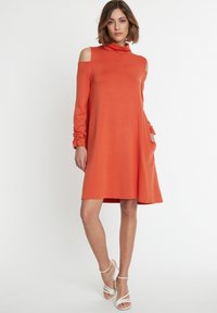 Orangekleid mit langen Ärmeln, hohem Ausschnitt, schulterfreien Ausschnitten und Seitentaschen. Glatte Textur, entspannte Passform und knielanger Saum.