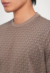 Brown t-shirt featuring a geometric pattern of overlapping "K" shapes. Round neckline and short sleeves, made of soft, textured fabric.