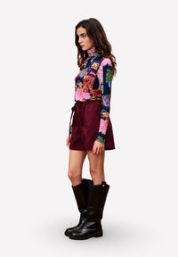 Een vrouw die zijwaarts staat in een kleurrijk gestreepte lange mouw top, bordeauxrode rok met strik, en zwarte kniehoge laarzen op een witte achtergrond.