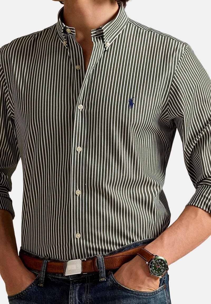 Chemise à rayures boutonnée en vert foncé et blanc, avec un col, arborant un petit logo, associée à une ceinture marron et une montre.