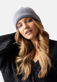 Szary wełniany beanie o prążkowanej strukturze, noszony lekko na bok. Model ma na sobie ciemny sweterek o teksturowanej powierzchni i ma jasnobrązowe faliste włosy.