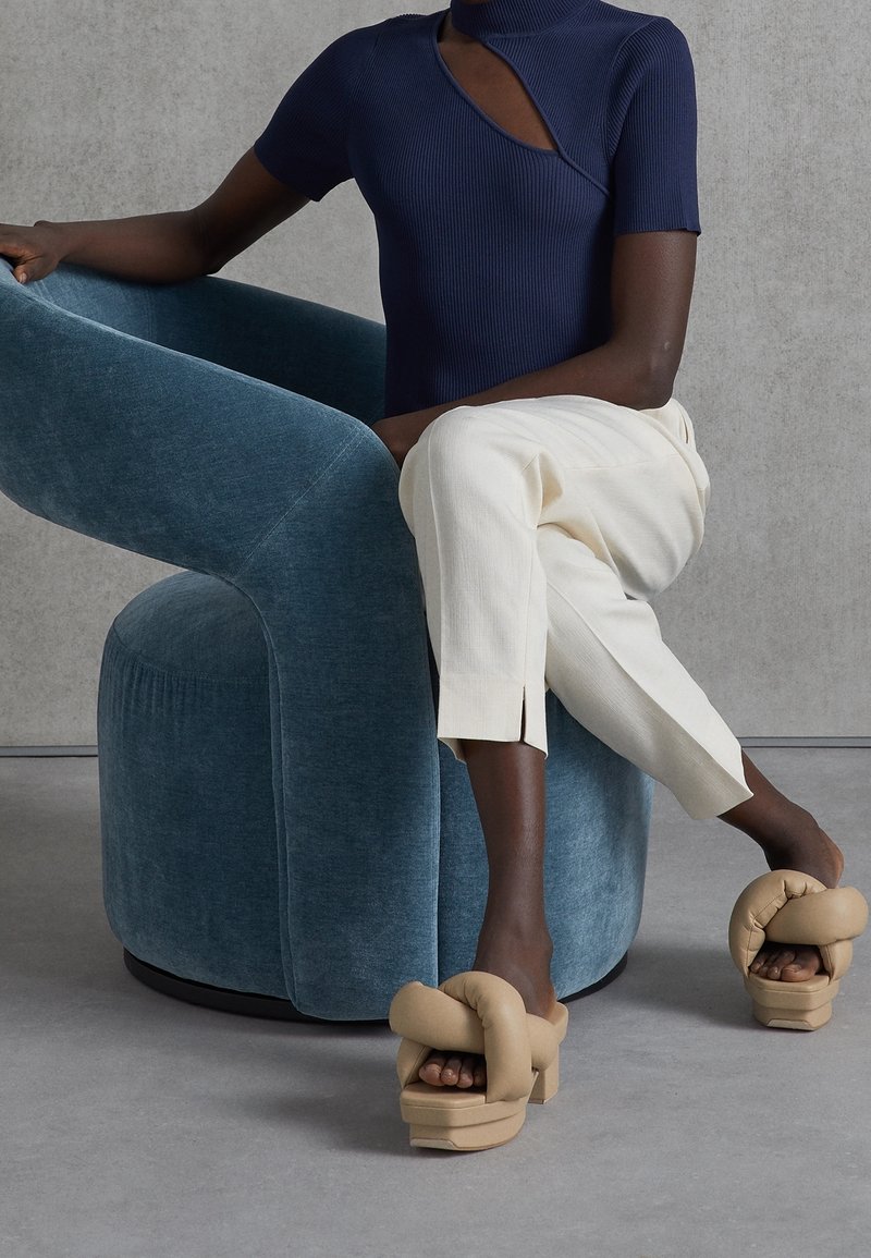 Beige vadderade plattformsandaler med en knutdetalj på remmen, i kombination med krämfärgade smala byxor och en mörk ribbad kortärmad topp, sittande på en blå stol.