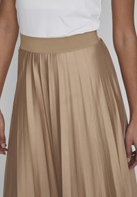 Jupe midi plissée beige, dotée d'une large ceinture élastique. Tissu lisse et brillant avec une texture douce et des plis verticaux définis.