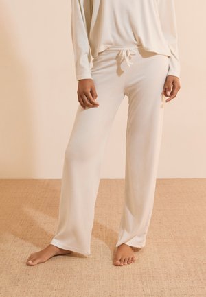 Personne debout pieds nus sur un tapis, portant un pantalon ample, clair avec cordon de serrage et un haut à manches longues assorti.