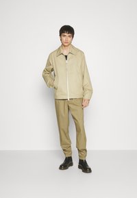 Beige jakke med synlig glidelås foran, parret med lysebrune bukser. Modellen har på seg svarte elastiske støvler. Glatt stofftekstur.