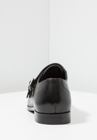 Scarpa da uomo in pelle nera con una fibbia in argento, fotografata dal retro su una superficie bianca contro uno sfondo grigio chiaro.
