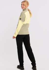 Chaqueta de forro polar verde claro y amarilla con un cuello alto, pantalones negros ajustados y zapatos negros. La chaqueta presenta un diseño a bloques de color y un logo.