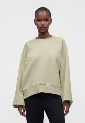 Person med et neutralt udtryk iført en løs, lysebeige sweatshirt og sorte bukser, stående mod en ensfarvet hvid baggrund.