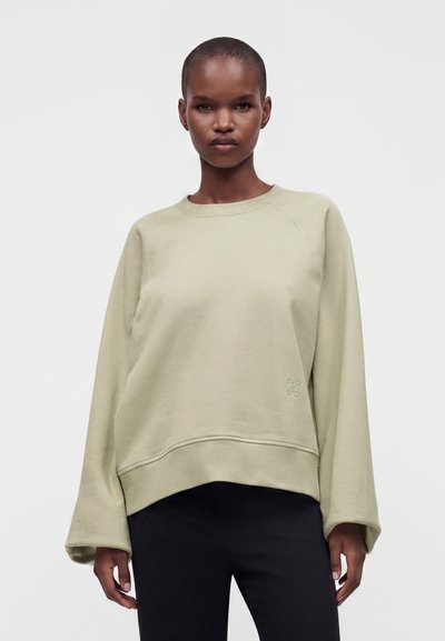 Personne avec une expression neutre porte un sweat-shirt beige clair ample et un pantalon noir, debout devant un fond blanc uni.