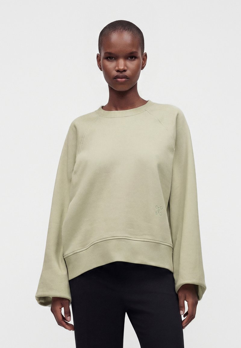Personne avec une expression neutre porte un sweat-shirt beige clair ample et un pantalon noir, debout devant un fond blanc uni.