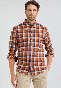 Man met baard die een oranje, blauw en witte geruite button-up shirt met opgerolde mouwen en beige broek draagt, staande tegen een effen achtergrond.
