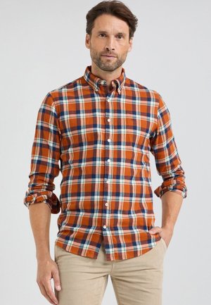 Man with beard wearing orange, blue, and white plaid button-up shirt with rolled sleeves and beige pants, standing against plain background.