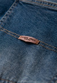 Denimstoff mit blauem Farbton und sichtbarer Textur. Ein braunes Lederetikett ist eingenäht, auf dem "ombre dept. seit 1986" steht.