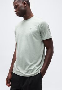 Νέος άνδρας που φοράει ανοιχτό πράσινο ριγέ μπλουζάκι Calvin Klein με τα χέρια στις τσέπες μαύρου παντελονιού, κοιτάζοντας προς τα κάτω και αριστερά.