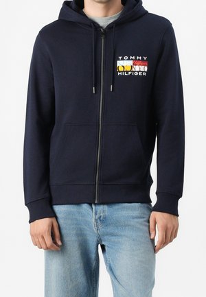 Uomo che indossa una felpa con cappuccio blu navy con cerniera e logo multicolore "Tommy Hilfiger" sul petto e jeans azzurri.