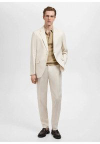 Lichtbeige pak met een enkelgeknoopte blazer, rechte broek en een tan ribbelshirt; gecombineerd met donkere, gestructureerde instapschoenen.
