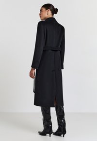 Cappotto lungo nero con vita accentuata da cintura, tessuto strutturato, colore noir e dettaglio con spacco sul retro, abbinato a stivali neri lucidi al ginocchio.