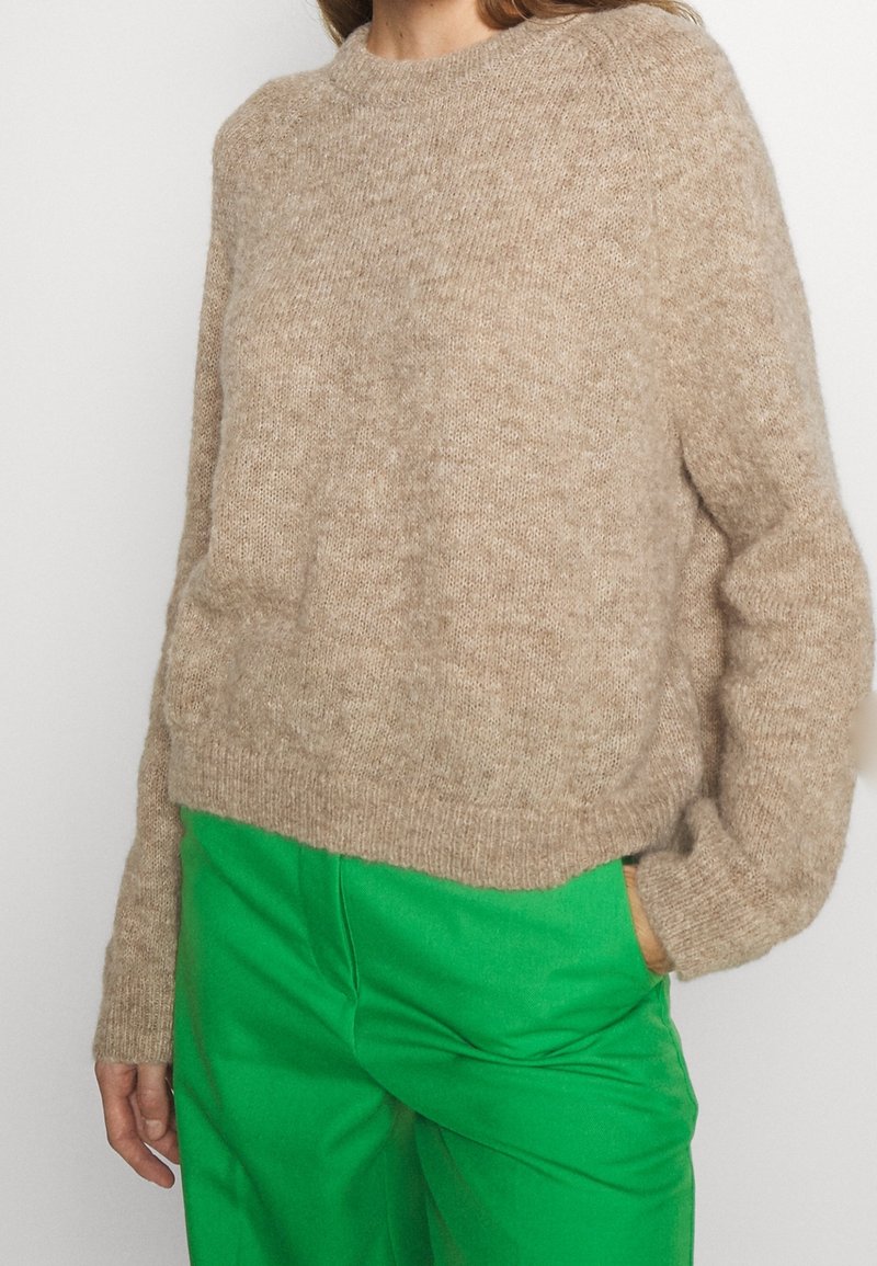Vrouw in een beige zachte gebreide trui en felgroene broek, met een hand in haar zak, tegen een plain witte achtergrond.