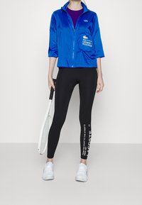 Osoba ve modré zipové bundě, černých legínách s bílým textem Lacoste, bílých teniskách, držící bílou tenisovou raketu po boku.