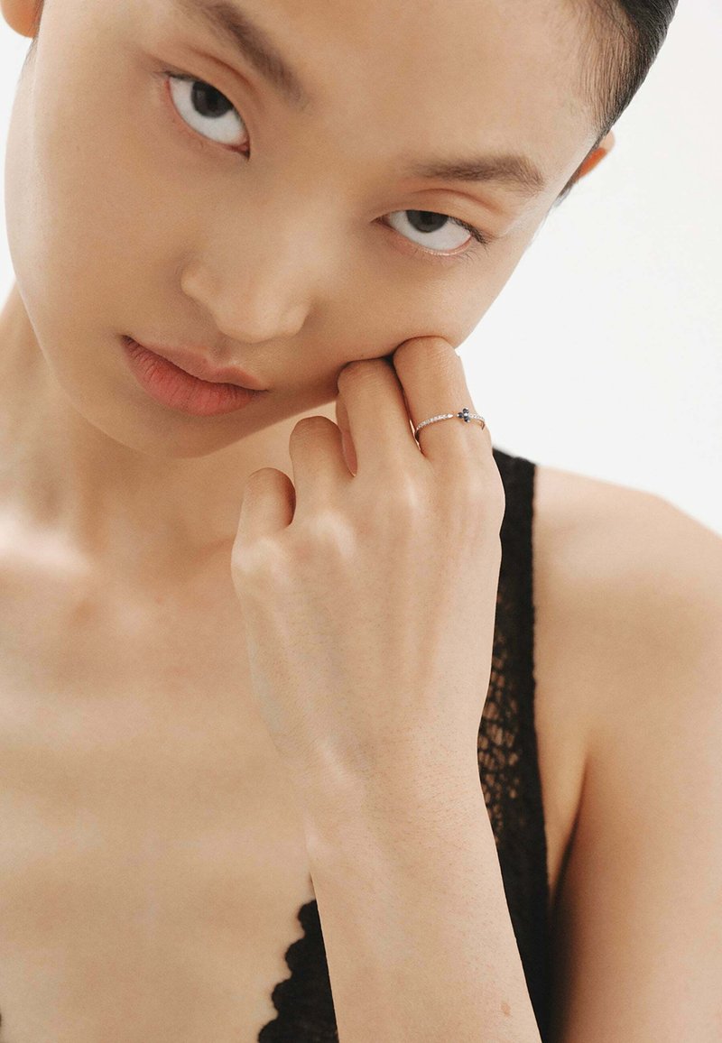Anillo de plata con una gema azul, con una bandita delicada y pequeñas piedras claras. La mano del modelo toca suavemente su rostro, con una textura de piel suave.