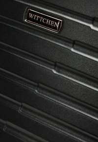 Superficie texturizada de maleta dura negra con relieves horizontales y una placa metálica rectangular con el logo grabado "WITTCHEN".
