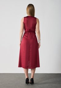 Burgundy satin klänning, utan ärmar, insvängd midja, midi längd med en slät textur. Stylerad med svarta högklackade skor.
