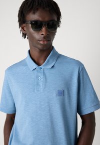 Polo de cor azul claro, feito de um tecido suave, apresenta um fecho de três botões e uma superfície texturizada com um pequeno logo no peito.