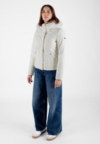 Giacca bianca imbottita con colletto in pelliccia sintetica, chiusura con zip e due tasche laterali, abbinata a jeans a gamba larga blu e sneakers bianche.