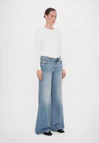 Bela dolga majica z dolgimi rokavi v kombinaciji s širokimi svetlo modrimi kavbojkami. Denim ima rahlo zbledelo barvo. Model nosi črne čevlje in stoji pred enobarvno ozadjem.