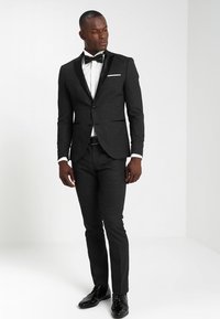 Schwarzer Anzug aus Wolle mit Satinakzenten, schlankes Design, mit Revers, weißem Hemd und Fliege, kombiniert mit glänzenden schwarzen Schuhen.