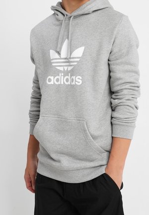 Person, die eine hellgraue Adidas-Kapuzenjacke mit weißem Logo und schwarze Hosen trägt, steht mit einer Hand in der Tasche vor einem einfarbigen Hintergrund.