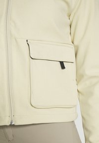 Beige jacka med en stor ficka fram och svart dragflik, bärs över ljusa byxor som visar dragkedja och sömmar.