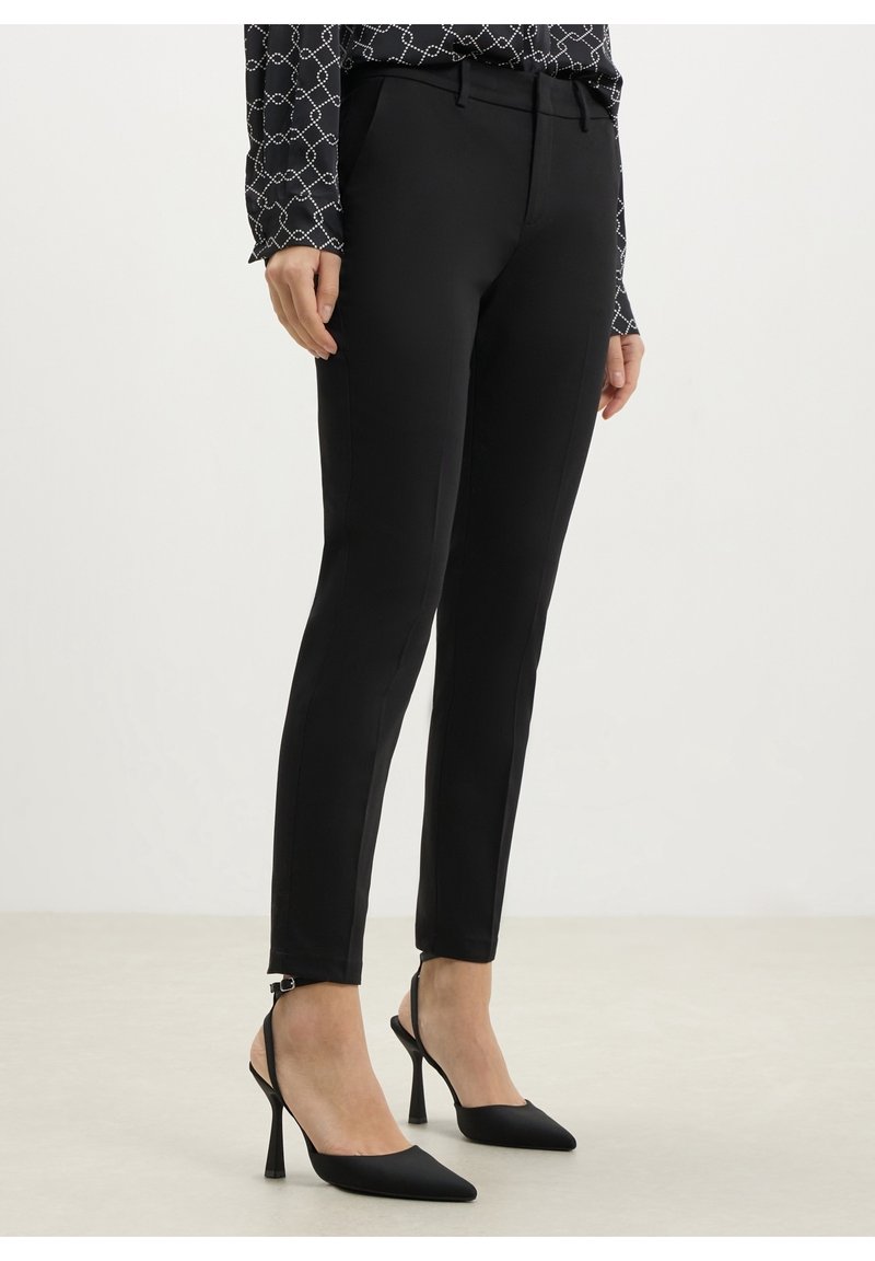 Femme portant un pantalon noir ajusté, un chemisier noir et blanc à motifs, et des chaussures slingback noires à talons hauts, debout sur un sol clair.