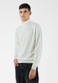 Sweter w kolorze kremowym z kołnierzem, wykonany z miękkiego materiału, z długimi rękawami i ściągaczami. Nosi się go z czarnymi dżinsami.