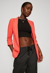 Blazer de coral con mangas enrolladas, combinado con un top corto negro y pantalones negros con cordón. Tejido suave y elementos de diseño ajustados.
